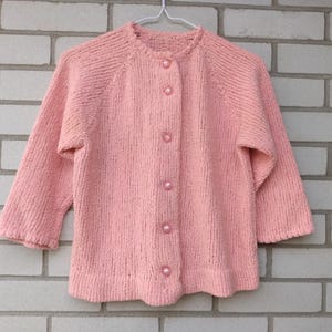 以下が含まれることがあります： 丸いネックラインと七分袖の、ヴィンテージスタイルのライトピンクのカーディガン。 セーターは、5つのマッチングピンクボタンが付いたボタンアップフロントを備えています。 ニットウェアは、レンガの壁を背景に白いハンガーに吊るされています。