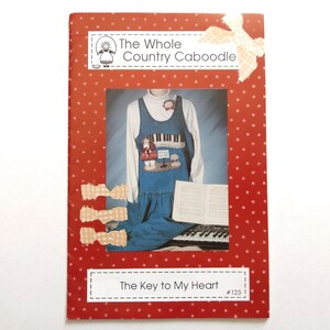 May include: A red card with white polka dots features a framed image of a denim jumper dress with a piano design. The text "The Key to My Heart" is displayed at the bottom. The card is decorated with paper bows.