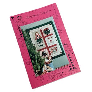 May include: A pink Christmas Sampler book with a decorative border. The cover features a cross-stitch design with a Christmas tree, a doll, a wreath, and the word "JOY".