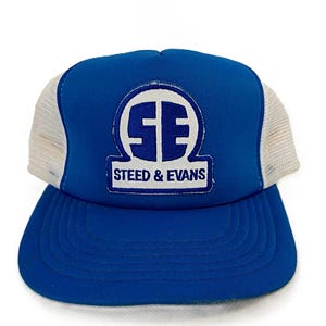 以下が含まれることがあります： 白のメッシュバックが付いた青いトラッカーハット。帽子の前面には、円の中に「SE」の文字が入った白いロゴがあり、その下に「STEED & EVANS」の文字が白で印刷されています。
