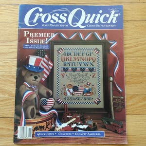 May include: The cover of a "Cross Quick" magazine features a patriotic theme. A teddy bear wearing a top hat and holding a heart-shaped flag is next to a framed cross-stitch sampler with an alphabet and patriotic motifs. The magazine's title is in blue.