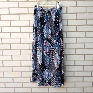 Peut inclure: Une jupe longue au sol présentant un motif patchwork avec divers imprimés floraux et géométriques dans des tons de noir, bleu, blanc et rose. La jupe possède une taille élastiquée avec un cordon de serrage et est suspendue à un cintre en plastique blanc.