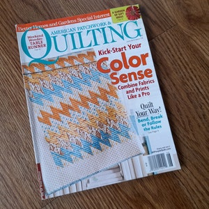 May include: A copy of "Quilting" magazine, featuring a quilt design in blue and orange zig-zag patterns. The cover promotes "Kick-Start Your Color Sense." The magazine rests on a wooden surface.