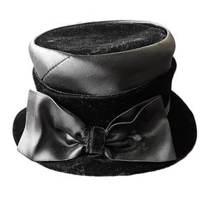 May include: A black velvet hat featuring a flat top and a structured crown. The hat is accented with a wide, smooth satin band around the base of the crown and a large, prominent satin bow attached to the front, resting on the brim.