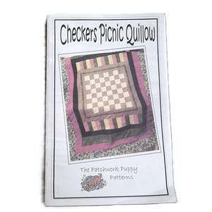 May include: A white paper pattern booklet titled Checkers Picnic Quillow by The Patchwork Puppy Patterns. The cover features a photograph of a quilt with a central checkerboard design in light and dark squares, surrounded by pink and black borders.