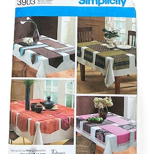 May include: A Simplicity sewing pattern envelope showing four table runner designs. The envelope is white with a blue banner and the number 3903. The designs feature diverse colours and patterns, with the name "Donna Babylon" visible.