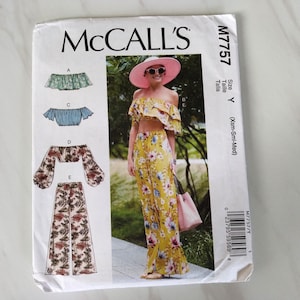 May include: McCall's M7757 sewing pattern for a two-piece outfit. The pattern includes a ruffled crop top and wide-leg pants with a floral print. The image shows a woman wearing the outfit, with a pink hat and purse.