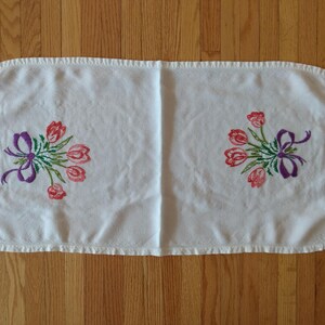 May include: White rectangular table runner with embroidered floral designs. The runner features two bouquets of red tulips with green leaves and purple bows. The edges are finished with a ruffled trim.