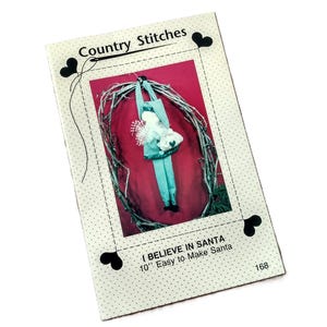 May include: A Country Stitches sewing pattern booklet titled I BELIEVE IN SANTA 10" Easy to Make Santa. The cover features a photograph of a fabric Santa doll with green and white checkered clothing, hanging inside a circular twig wreath.