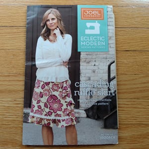 May include: A sewing pattern booklet for a cascading ruffle skirt by Joel Dewberry. The cover features a person wearing a white long-sleeve top and a cream skirt with a pink and dark red floral pattern. Text includes Eclectic Modern Sewing Patterns.