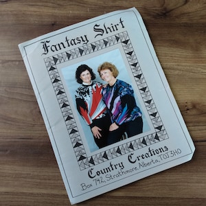 Peut inclure: Une pochette de patron en papier intitulée Fantasy Shirt par Country Creations, présentant une photographie de deux personnes portant des chemises à manches longues à motifs. La bordure a un motif géométrique noir et blanc.