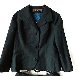 May include: A dark green suit comprising a blazer and skirt. The blazer features a notched collar, three buttons, and a teal lining. The suit is displayed on a hanger against a white backdrop.