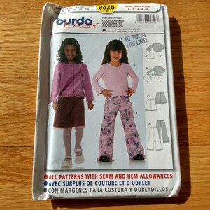 以下が含まれることがあります： Burda Easyの型紙封筒、番号9826。長袖トップス、スカート、ワイドパンツを着用した子供たちのイラストが描かれています。パッケージには多言語のテキストと、衣類のパーツの図面が含まれています。