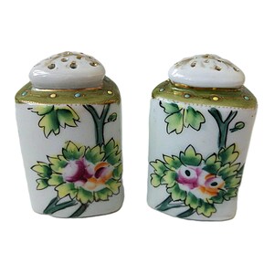 May include: A pair of antique salt and pepper shakers. The shakers are white with a floral design featuring pink, yellow, and green flowers. The tops are white with small holes and a green and gold trim.