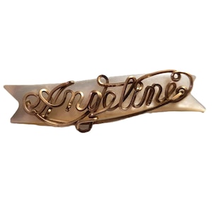 Peut inclure: Une barrette à cheveux vintage avec le nom "Angeline" en fil d'or élégant. L'écriture est fixée sur une base blanche crème en forme de bannière. La barrette est un accessoire de cheveux décoratif.