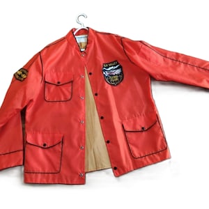 May include: A bright orange jacket with a tan lining, featuring a snap-button front closure and black piping trim. The chest displays a patch reading Elk Valley Flying Club, while a smaller patch is on the right sleeve. It has four front pockets.