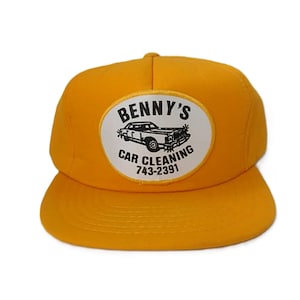 May include: A bright yellow baseball cap featuring a white oval patch on the front. The patch displays a black graphic of a car with the text BENNY'S CAR CLEANING and the phone number 743-2391 printed in black lettering across the centre.