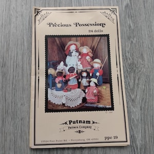 May include: A vintage pattern booklet titled "Precious Possessions" featuring a collection of 24 dolls. The cover displays a framed image of the dolls, with the Putnam Pattern Company logo and address at the bottom. The booklet is on a wooden surface.