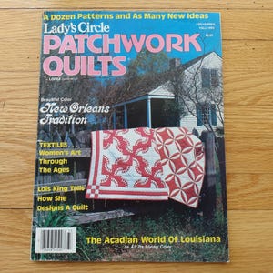 May include: The cover of Lady's Circle Patchwork Quilts magazine, showcasing a red and white quilt over a fence. Text on the cover mentions quilting patterns, the New Orleans tradition, and the Acadian World of Louisiana.