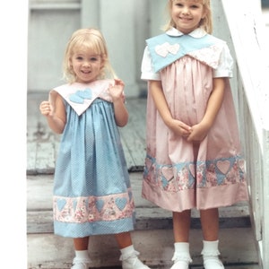 May include: Two young girls wearing handmade dresses. One dress is light blue with white polka dots, the other is pink. Both dresses have heart-shaped accents and floral trim at the hem. The girls are standing on a wooden staircase.