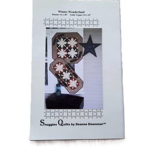 May include: A printed pattern for a winter-themed quilt project. The design includes a runner (14 x 30 inches) and a table topper (24 x 24 inches). The image shows the finished quilt designs with star and floral motifs.