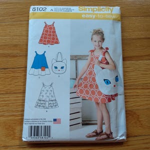 May include: A Simplicity 8102 easy-to-sew pattern envelope featuring illustrations of three different dress styles and a cat-shaped bag. The cover displays a photograph of a child wearing the orange circle-patterned dress and carrying the white cat bag.