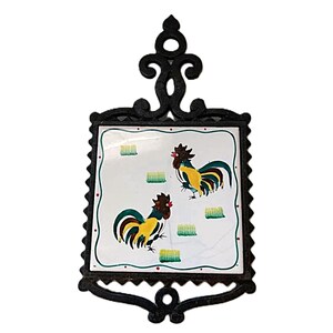 May include: A black cast iron trivet featuring a square white ceramic tile insert. The tile displays two roosters with yellow, green, and red plumage, surrounded by small green rectangular shapes and red dots along the border of the tile.