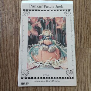May include: A pattern titled "Punkin' Patch Jack" featuring a pumpkin-shaped scarecrow with a straw hat and plaid accents. The image is 15 cm tall and includes the text "Homespun at Heart Designs".