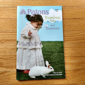 May include: A Patons knitting pattern booklet titled "Bamboo Baby and Bunnies" featuring a young child in a white dress and a white rabbit on green grass. The booklet includes instructions for knitting.