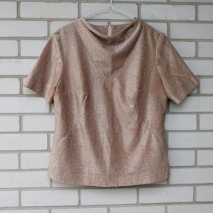 May include: A short-sleeved, tan-coloured top with a textured, shimmering fabric. The top features a unique neckline and a tailored fit, hanging on a white hanger. The garment is displayed against a white brick wall.