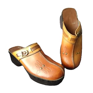 May include: A pair of brown leather clogs with a buckle and decorative stitching. The clogs have a platform sole and a wooden heel. The leather is a light brown colour with darker brown accents. The clogs are in good condition and appear to be vintage.
