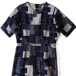 May include: A short-sleeved dress featuring a geometric patchwork pattern in shades of navy blue, slate grey, and light grey. The garment has a round neckline, a fitted waistline, and a visible black zip closure at the centre back of the neck.