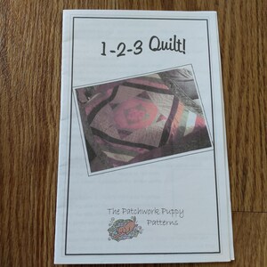 May include: A white instructional booklet titled 1-2-3 Quilt! featuring a central photograph of a multi-coloured geometric quilt. The bottom of the cover displays the text The Patchwork Puppy Patterns with a small illustration of a brown puppy.