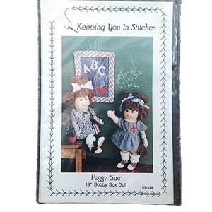 May include: A package for a Peggy Sue 13-inch Bobby Sox Doll. The package features two dolls with brown hair and dresses, standing in front of a chalkboard with the text "ABC" and "I love Bobby Joe". The package is labeled "Keeping You In Stitches".