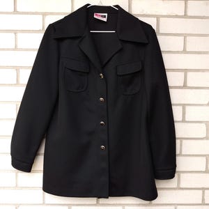 May include: A black vintage jacket with a collared neckline, two front pockets, and gold-colored buttons. The jacket is hanging on a white hanger against a white brick wall.