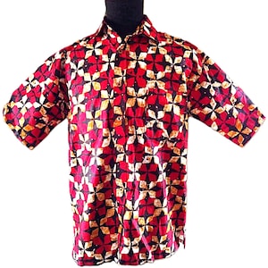 May include: A short-sleeve button-down shirt featuring a repeating geometric pattern of four-pointed stars in shades of red, black, and cream. The shirt has a standard collar, a front button placket, and a straight hemline with a consistent print.