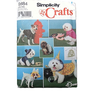 May include: A Simplicity Crafts 9884 sewing pattern envelope featuring illustrations of dogs wearing various costumes including a cape, hat, and shirt. The white paper packaging displays multiple canine models in different outfits against a green background.