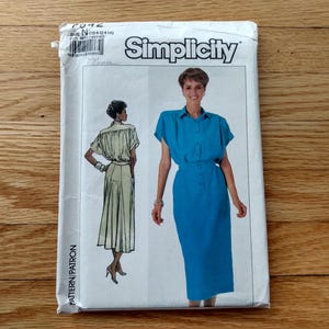 May include: A Simplicity sewing pattern packet featuring illustrations of a woman in a blue dress and a woman in a beige outfit. The packet has the Simplicity logo and the text "PATTERN/PATRON". The packet is on a wooden surface.