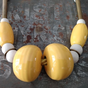 Peut inclure: Gros plan sur un collier composé de grosses perles jaunes brillantes et de petites perles blanches. Le collier a une esthétique vintage, avec des accents en métal couleur laiton et un fond neutre.