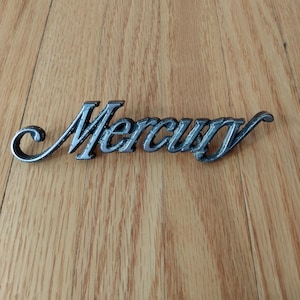 May include: A chrome-coloured Mercury emblem, displayed on a wooden surface. The emblem is in a cursive font, with the word "Mercury" spelled out. The emblem is likely from a vintage car.
