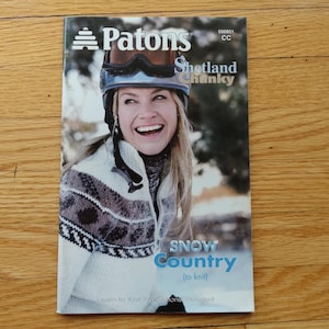 May include: A Patons Shetland Chunky knitting pattern booklet titled Snow Country. The cover features a smiling person wearing a white, grey, and brown patterned sweater and a ski helmet with goggles. The text includes Patons, Shetland Chunky, and Snow Country.
