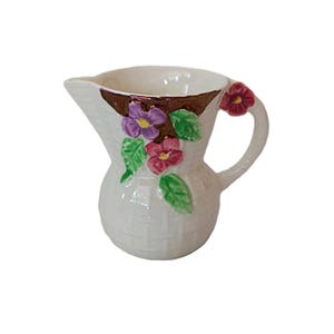 May include: A white ceramic pitcher with a woven basket design. The pitcher features purple and red floral accents with green leaves. The rim is brown, and the handle has a red flower. A decorative piece for serving.