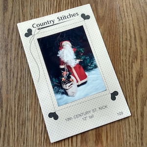 May include: A rectangular paper pattern envelope titled Country Stitches featuring a photograph of a Santa Claus doll with a white beard, red coat, and a sack of greenery. The text reads 19th. CENTURY ST. NICK, 12" tall, and 109. The border has heart icons.