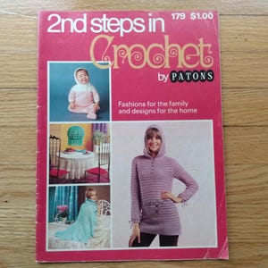 May include: A vintage crochet pattern book titled "2nd steps in Crochet by PATONS." The cover is pink and features images of crochet projects, including a baby in a pink outfit and a woman in a hooded sweater. The book promises fashions for the family and home designs.
