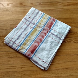 Peut inclure: Un torchon de cuisine blanc plié présentant un motif à carreaux avec de fines lignes rouges et bleues, accentué par une bande verticale de motifs circulaires jaunes et rouges. Le tissu est blanc avec un design en grille formé par les lignes.