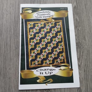 May include: A rectangular quilt pattern booklet titled Absolutely Positively Quilt Designs Change It Up, featuring a dark blue, gold, and green geometric chevron pattern on the cover with gold ribbon graphics and the code APQD 134.