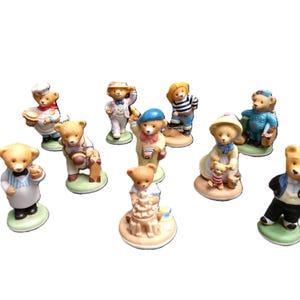 May include: A collection of nine small ceramic bear figurines, each depicted in different outfits and activities, including a chef, a traveller with a suitcase, a photographer, a sailor, a parent with a child, and figures in formal attire.