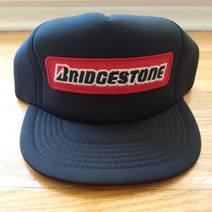 May include: Black trucker cap with a curved peak. The cap has a red rectangular patch with the white "BRIDGESTONE" logo. The hat is made of a sturdy material and is suitable for outdoor activities.