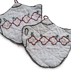 May include: Two white quilted fabric pot holders shaped like teapots. Each features a black stitched outline, a small red hanging loop, and a decorative row of embroidered red flowers with green leaves and yellow centers across the front surface.