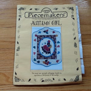 May include: A vintage "Piecemakers" pattern titled "Autumn Girl" featuring a hand-appliqued wall hanging design. The pattern includes a central oval image of a girl in a dress, surrounded by a floral border, and is on a cream-coloured background.